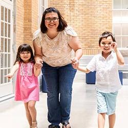 A smiling mother holding hands with her two children, walking through a bright hallway with large windows. The children are happy, with one child in a white shirt and blue pants, and the other in a pink dress.